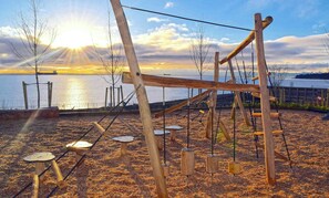 Un ensemble de jeux composé de balançoires en bois et de structures d’escalade, installé sur une zone sablonneuse, avec une vue sur le coucher de soleil au-dessus de l’eau.