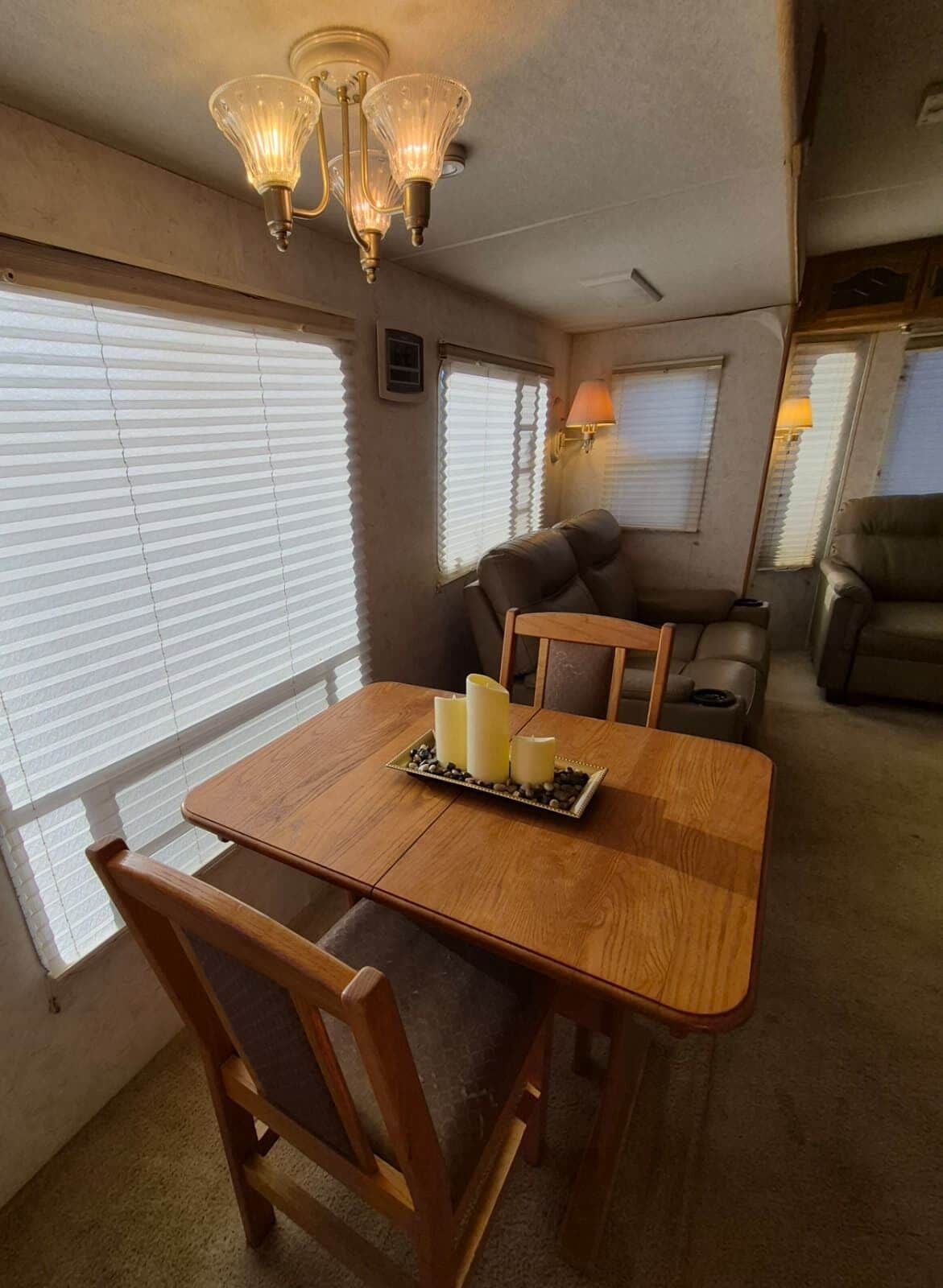 A dining area with a wooden table, chairs, and a chandelier. There are windows with blinds, a sofa, and a lamp.