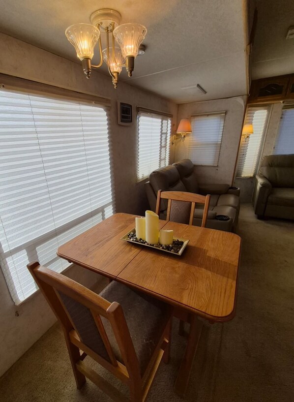 A dining area with a wooden table, chairs, and a chandelier. There are windows with blinds, a sofa, and a lamp.
