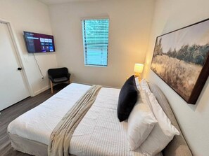 Habitación de hotel con cama, televisión montada en la pared, ventana con persianas y un cuadro en la pared.