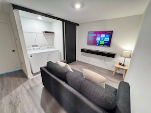Ein modernes Wohnzimmer mit grauem Sofa, einem flachbildfernseher an der Wand und einem Waschbereich mit Waschmaschine und Trockner.