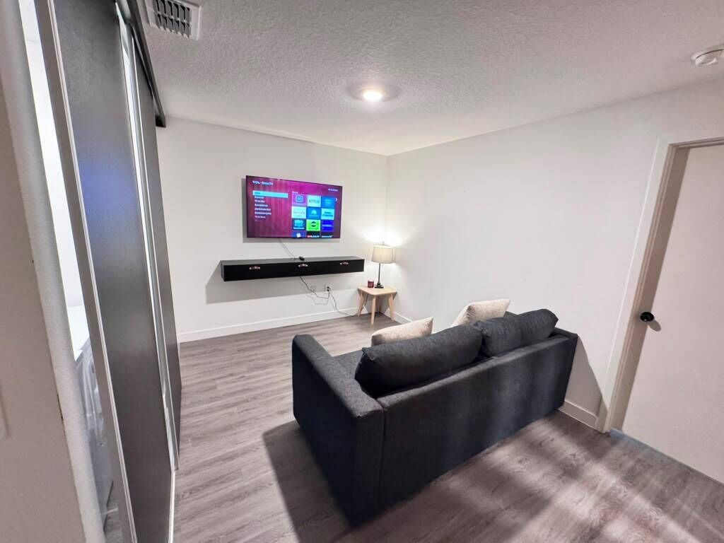 Una sala de estar moderna con un sofá oscuro, un televisor montado y una mesita de noche con una lámpara.