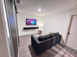 Una sala de estar moderna con un sofá oscuro, un televisor montado y una mesita de noche con una lámpara.