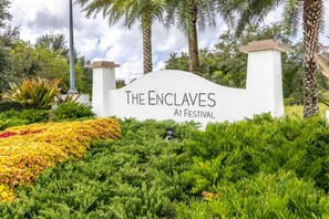 Ein Schild für 'The Enclaves at Festival' mit weißem Hintergrund und schwarzer Schrift, umgeben von üppigem Grün und Palmen.