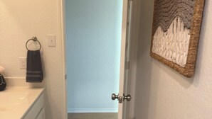 Ein Badezimmer mit einem weißen Waschbecken, einem Handtuchhalter, einem dunklen Teppich und einer Tür zu einem Raum mit hellblauer Wand und Holzboden.