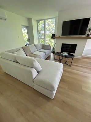 Ein Wohnzimmer mit einem Ecksofa, einem Flachbildfernseher über einem Kamin und einem kleinen Tisch mit Lampe.