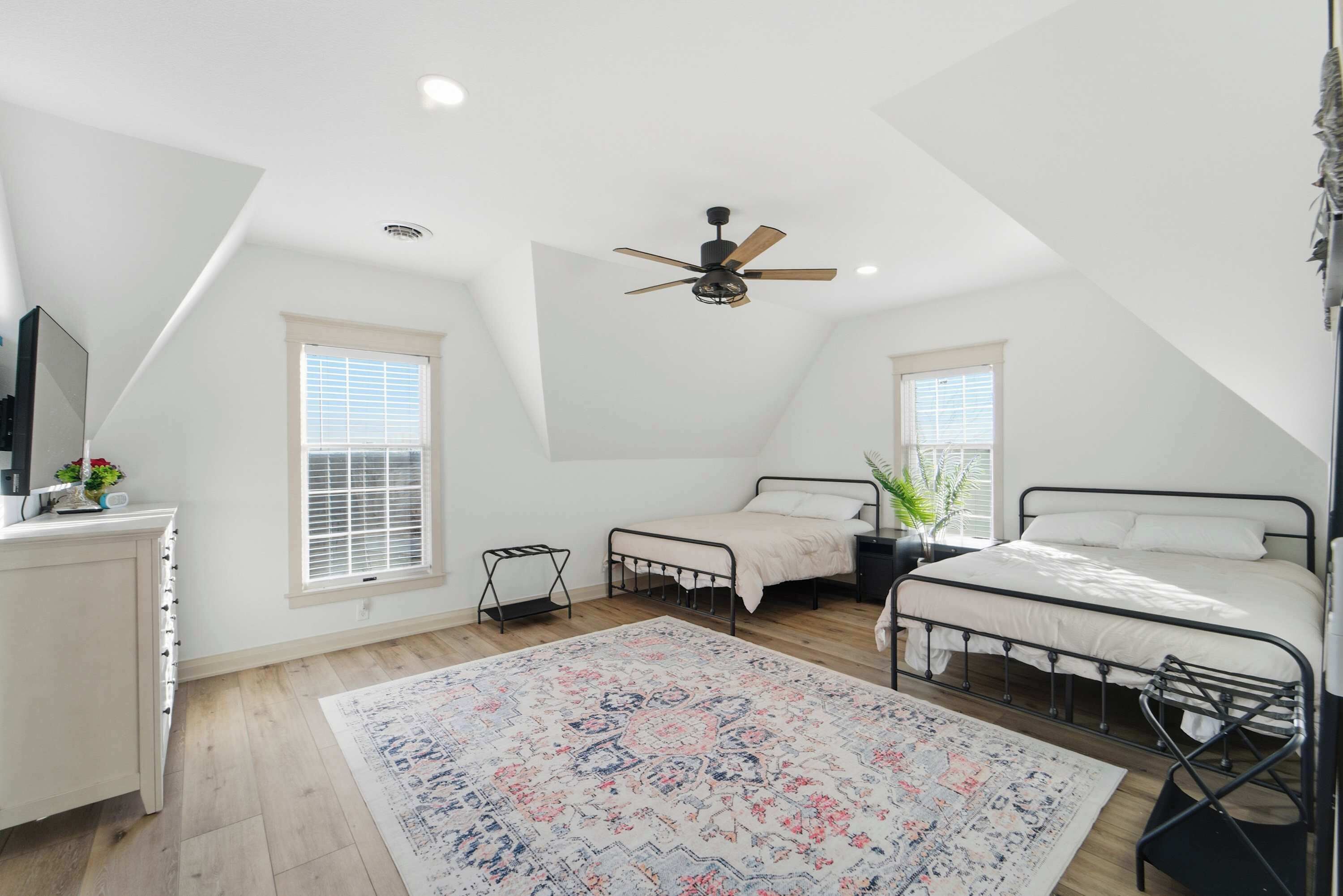A bedroom with a sloped ceiling, a ceiling fan, two beds, a bedside table, a nightstand, a window, a radiator, and a patterned rug.