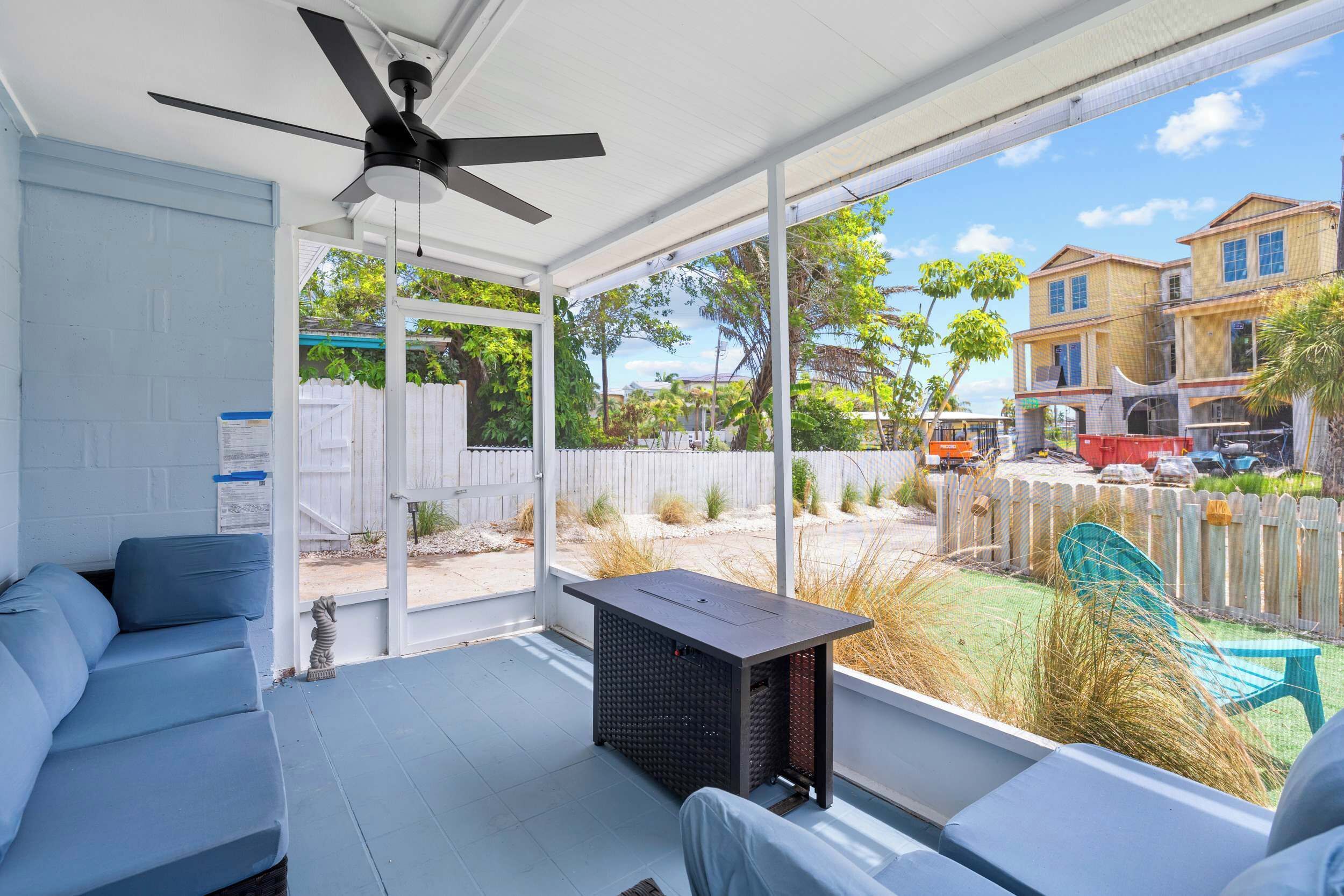 A covered patio with a ceiling fan, a seating area with a sofa, and a view of a residential area with houses and a fence.