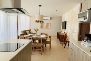 A modern kitchen with a built-in stove, dining area with wooden chairs, a hanging light fixture, and a flat-screen TV mounted on the wall.