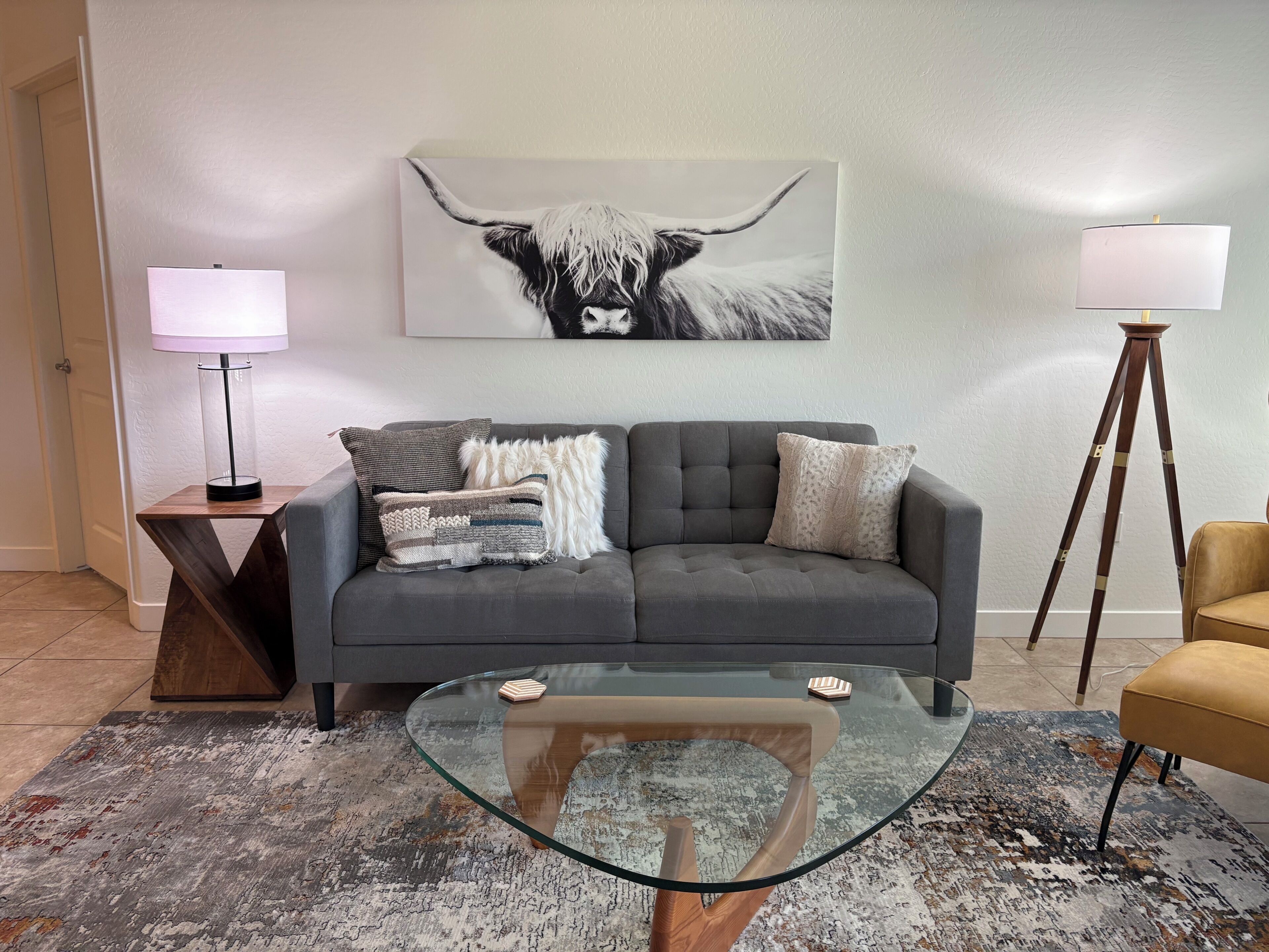 En moderne stue med en grå sofa, et glasbord, et mønstret tæppe, en lampe og et maleri af en tyr.