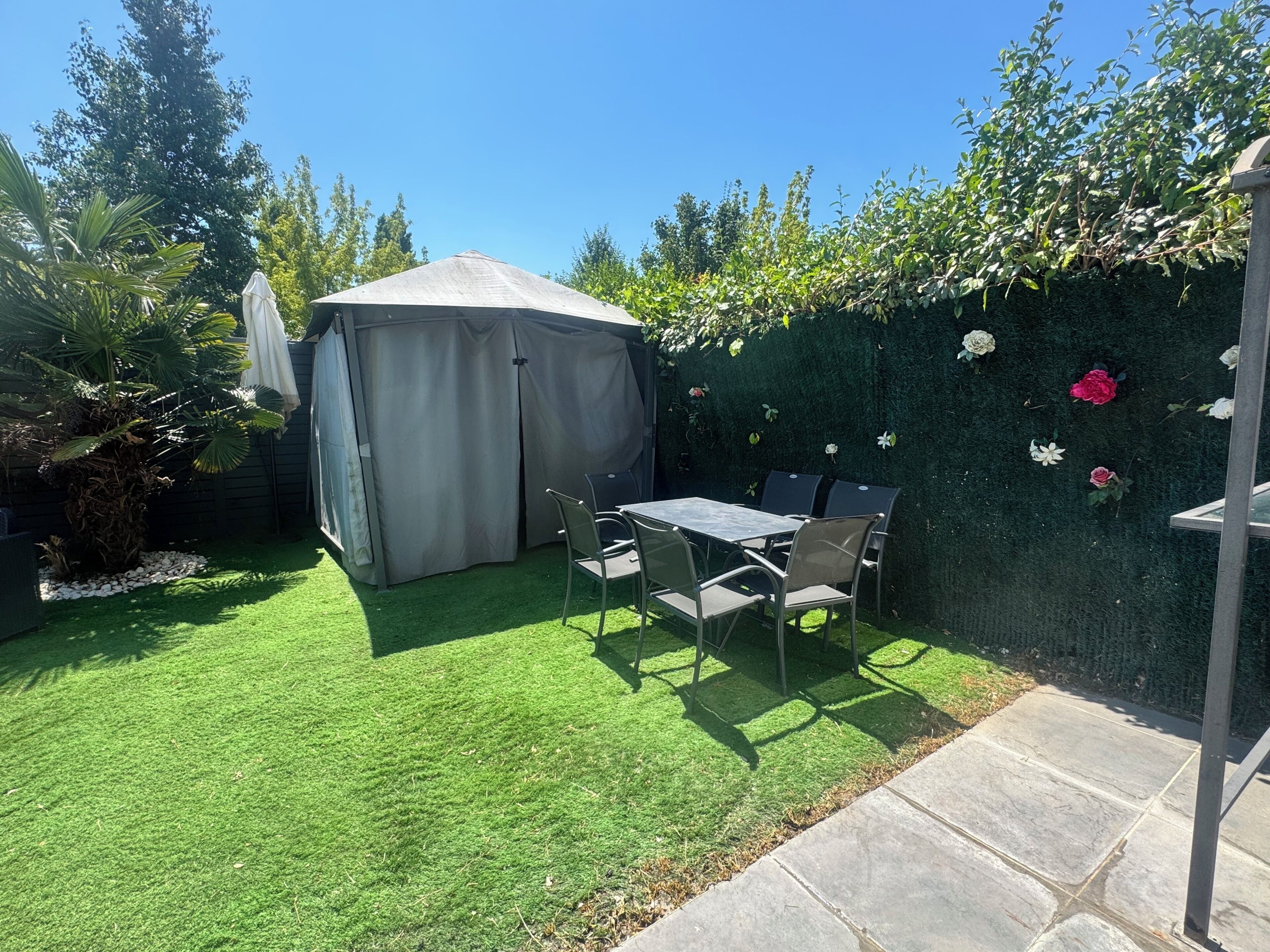 Eine Terrasse mit Tisch und Stühlen, ein überdachter Bereich und ein Blumenhecke.