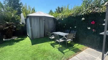 Eine Terrasse mit Tisch und Stühlen, ein überdachter Bereich und ein Blumenhecke.