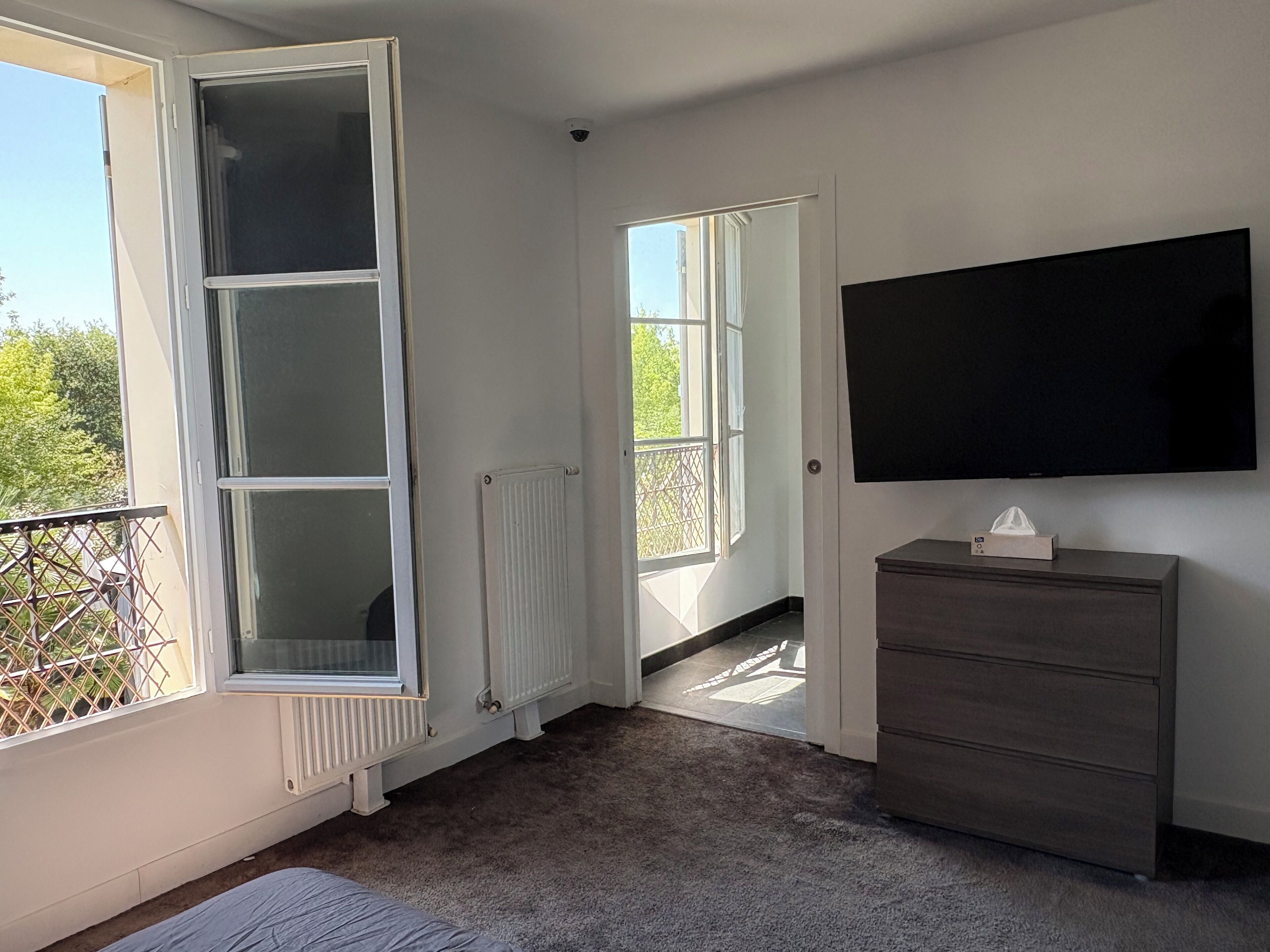 A room with a large window, a radiator, a flat-screen TV, and a dark wooden dresser.