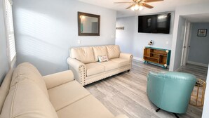 A living room with a beige sofa, a teal armchair, a wooden cabinet with a TV above it, and a ceiling fan. - Beautiful spacious upgraded 2-bedroom condo sleeps 8 (St. Pete Beach)