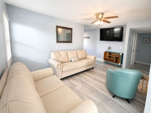 Ein Wohnzimmer mit einem beigefarbenen Sofa, einem türkisfarbenen Sessel, einer Holzkommode mit einem Fernseher darüber und einem Deckenventilator.