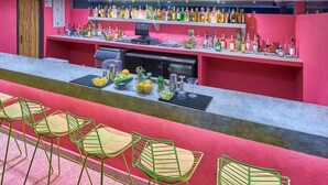Angolo bar con una palette di colori rossi e rosa, che include diverse bottiglie di liquore, una cassa registratrice e attrezzi da bar. Sono presenti sedie verdi e un bancone con frutta e verdura fresca.