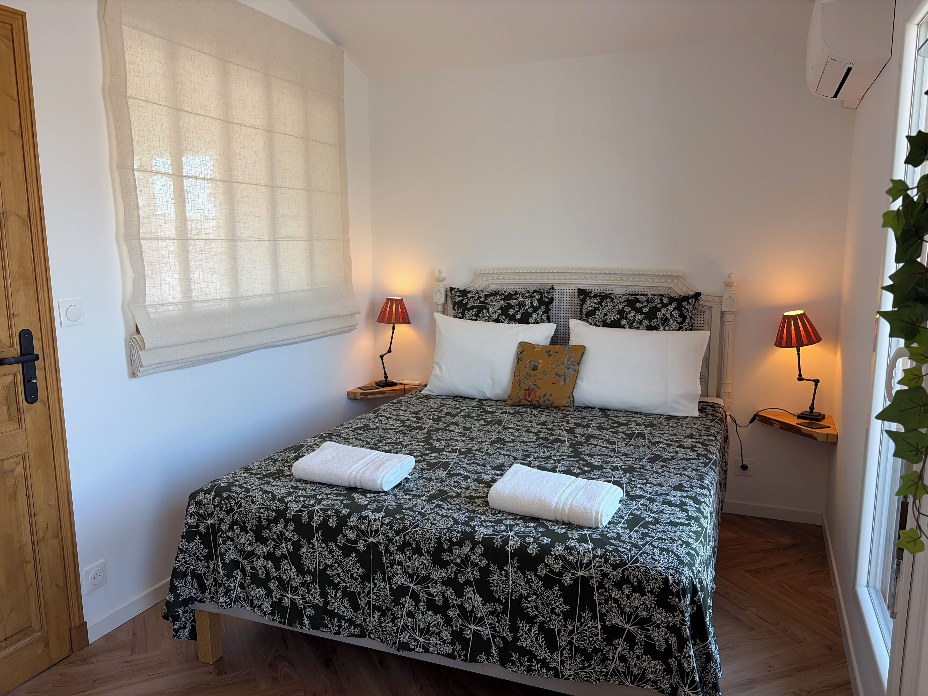 Una cama bien hecha con ropa de cama floral, dos lámparas de noche, un ventanal con cortinas translúcidas y una puerta de madera.
