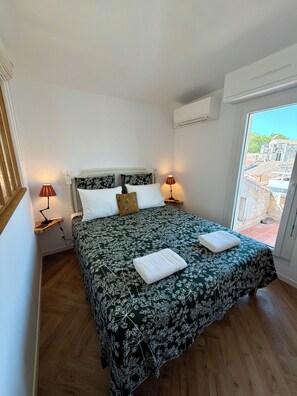 Ein Schlafzimmer mit einem großen Bett, zwei Nachttischlampen, einem Fenster mit Blick auf Gebäude und einer Klimaanlage.