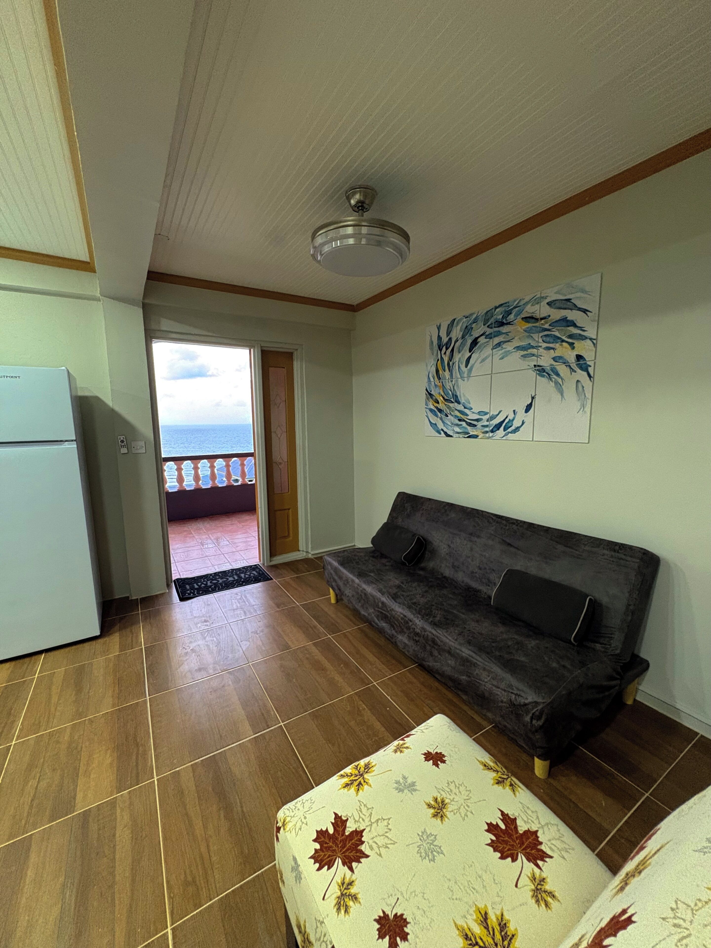 Una sala de estar con sofá, refrigerador, puerta que da a un balcón y ventilador de techo.