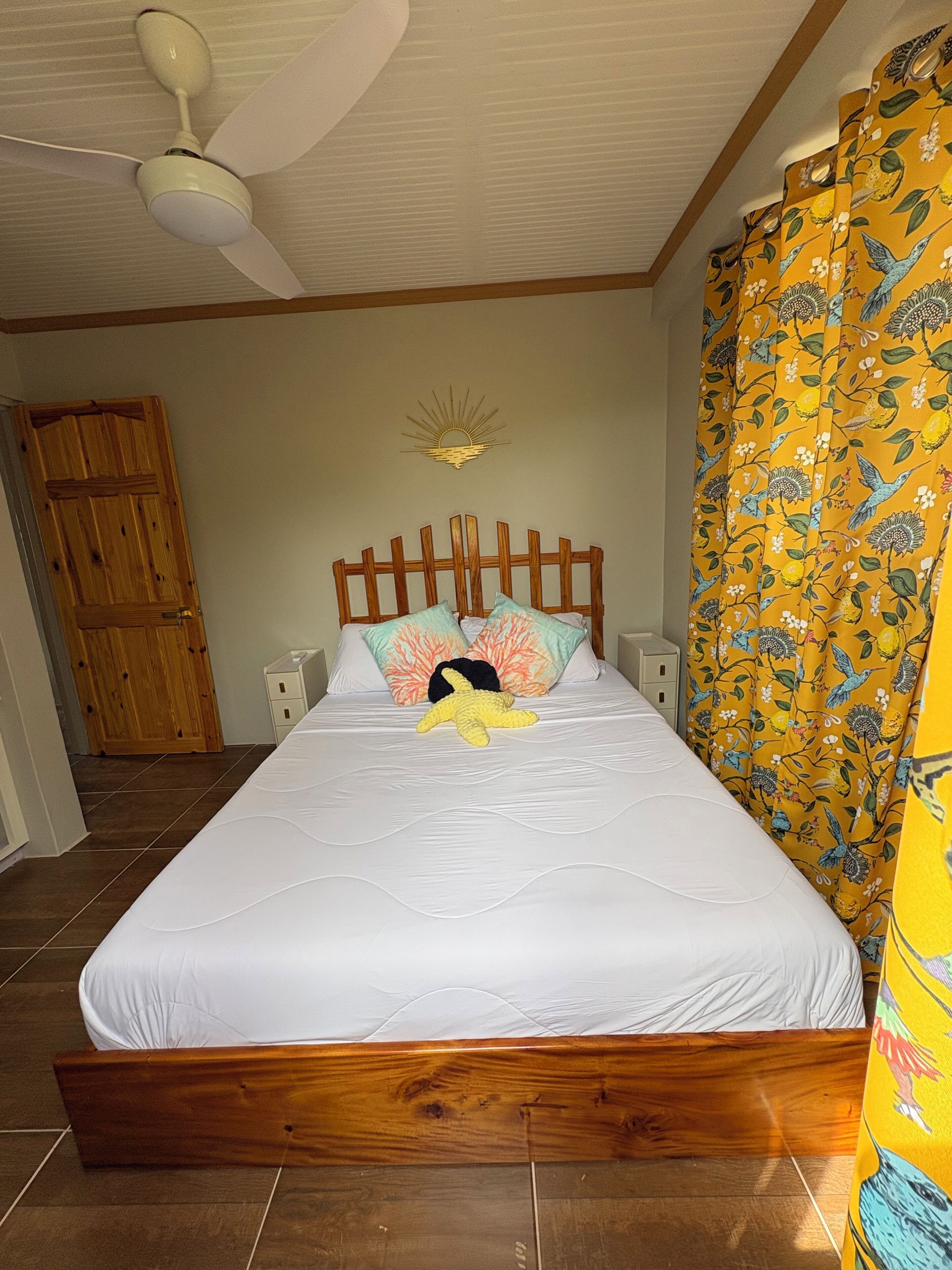 Een slaapkamer met een houten bed, wit beddengoed, een plafondventilator, een decoratief wandobject en een houten deur.