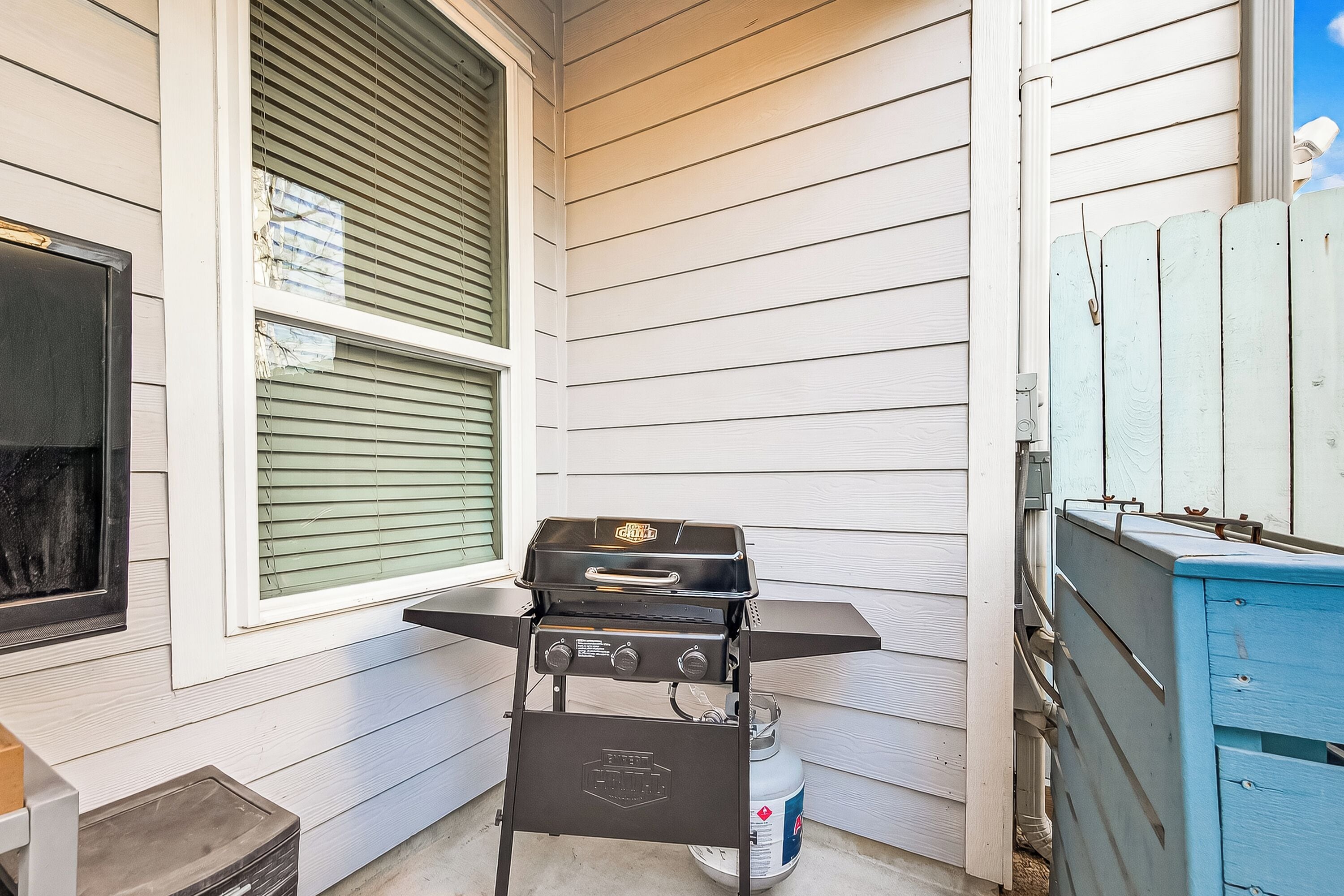 A small outdoor area with a grill and propane tank, adjacent to a house with white siding and a window with blinds.