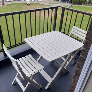 A balcony with a white table and two white chairs, a black railing, and a view of a grassy area with cars and buildings.