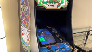 En arkadmaskin i retrostil med en färgstark design, utrustad med en joystick och knappar, och en skärm som visar olika spelalternativ.