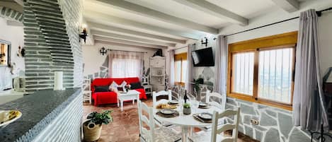 A spacious living area with a stone wall, a red sofa, a dining table set for four, and a television mounted on the wall.