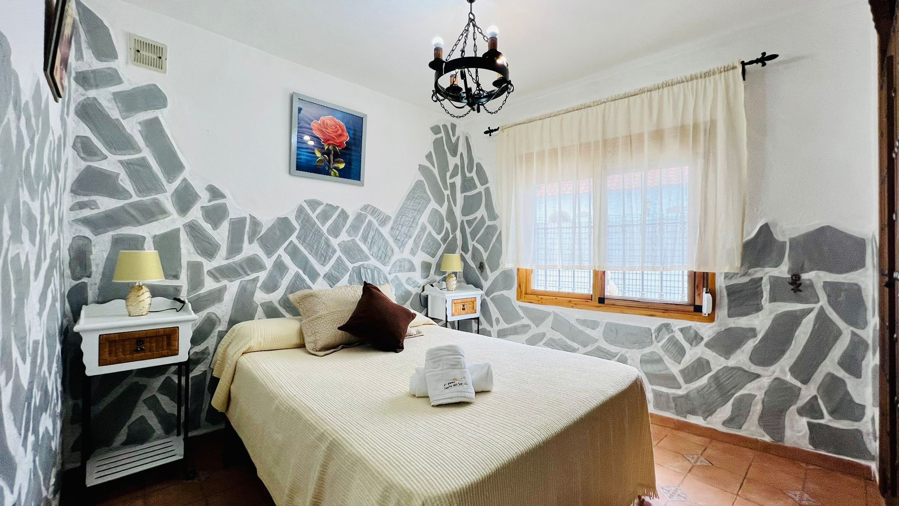 A bedroom with a stone-patterned wall, a bed with a white cover, two bedside tables with lamps, a window with curtains, and a chandelier.