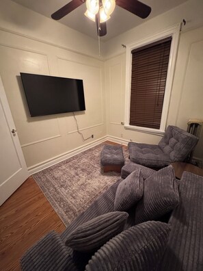 A room with a flat-screen TV mounted on the wall, a ceiling fan, a window with blinds, a patterned rug, and a sectional sofa. - Quiet Old Brooklyn Charm Upstairs 2-bedroom/1-bath Unit in E. Flatbush Townhome (Brooklyn)