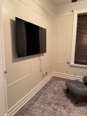 A room with a mounted flat-screen TV, a patterned carpet, a window with blinds, and a door with a glass panel. - Quiet Old Brooklyn Charm Upstairs 2-bedroom/1-bath Unit in E. Flatbush Townhome (Brooklyn)