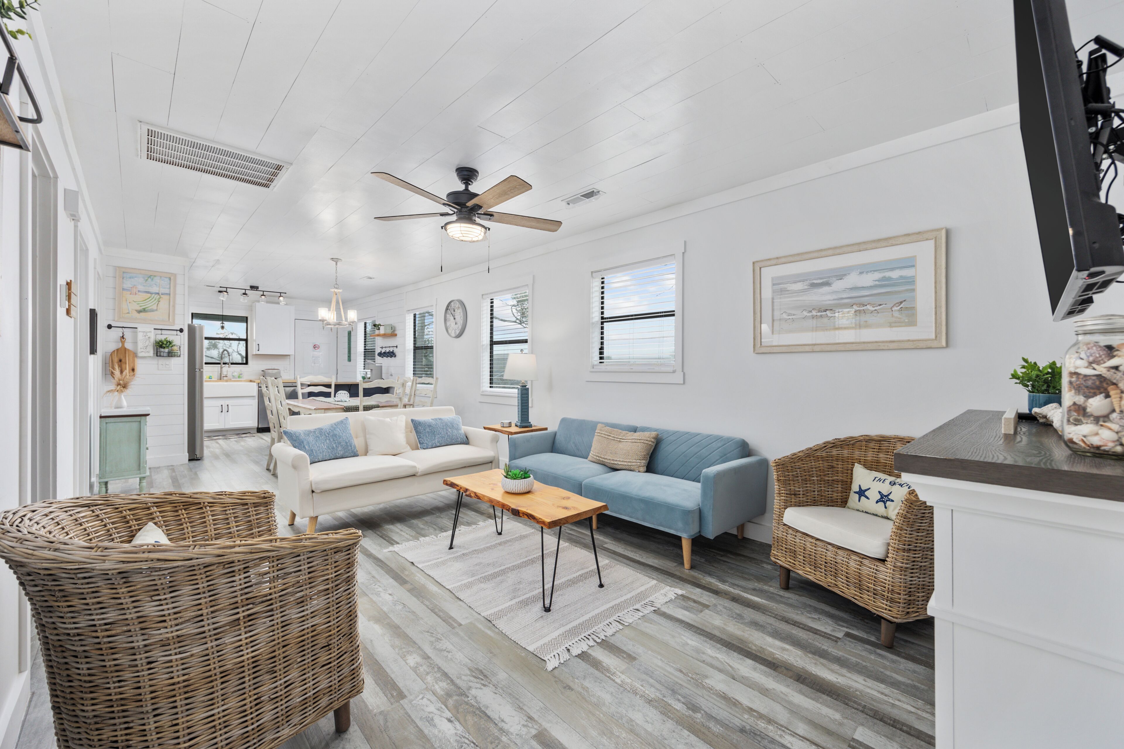 A spacious living room with a white sofa, blue sofa, wicker chairs, a wooden coffee table, and a ceiling fan.