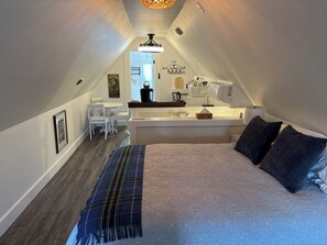 A bedroom with a bed, pillows, a bedside table, and a view into the adjacent kitchen area.
