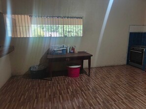 A room with a wooden floor, a window with blinds, a small table with items, a red bucket, and a stove. -  5000 Square feet house for rent. (Ticuantepe)