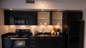 Une cuisine moderne avec des meubles de couleur noire, un four encastré, un four à micro-ondes, un réfrigérateur et un meuble de rangement avec des étagères en verre, où sont exposées des tasses et des bols.