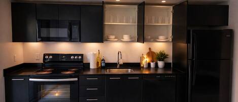 A modern kitchen with black cabinetry, a built-in oven, a microwave, a refrigerator, and a glass-fronted shelving unit with cups and bowls.