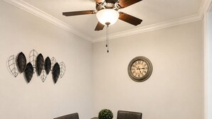 Een eetkamer met een houten tafel, zes stoelen, een plafondventilator, een wandklok en decoratieve wanddecoraties.