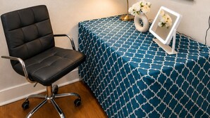 Een kamer met een zwarte bureaustoel, een tafel met een blauwe tafelkleed met dessin, een wandlamp en een lijst met bloemen.