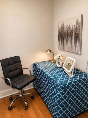 Ein Zimmer mit schwarzem Bürostuhl, einem Tisch mit blau kariertem Tischläufer, einer Wandlampe und einem Bilderrahmen mit Blumen.