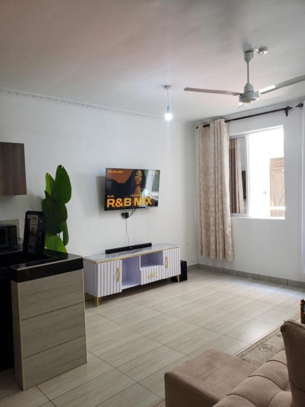 A living room with a flat-screen TV mounted on the wall, a ceiling fan, a window with curtains, and a sofa.