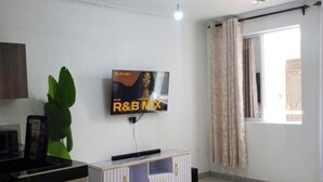 A living room with a flat-screen TV mounted on the wall, a ceiling fan, a window with curtains, and a sofa. - SPACIOUS VIP 1 BEDROOM with ROOFTOP SWIMMING POOL AVAILABLE (Mtwapa)