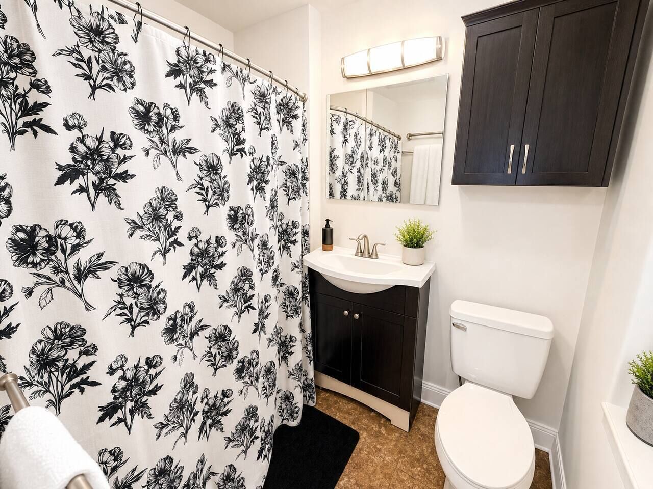 Een badkamer met douchegordijn met bloemenprint, een wit wastafelmeubel met spiegel erboven, een donkere kast en een wit toilet.