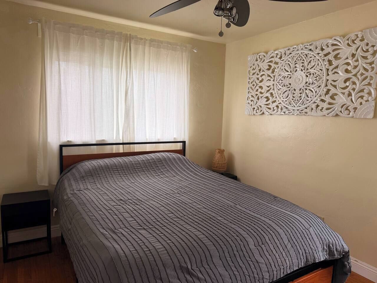 A bedroom with a bed, a nightstand, a ceiling fan, a window with curtains, and a decorative wall panel.