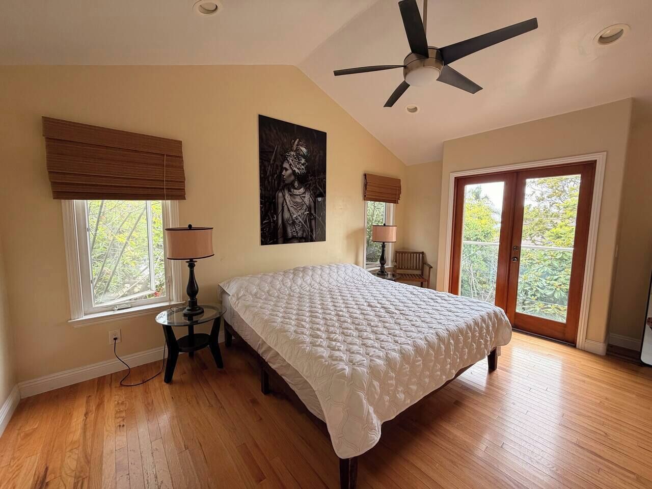 A bedroom with a bed, a ceiling fan, a window with blinds, a lamp, and a door leading to an outdoor area.