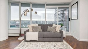 Una sala moderna con un sofá gris, alfombra estampada, lámpara de pie y ventanas amplias con vistas a un paisaje urbano.