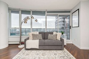 A modern living room with a gray sofa, patterned rug, floor lamp, and large windows overlooking a cityscape. - Perfect Vancouver Getaway Central 2BR/2BA+Sofa Bed (Vancouver)
