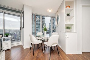 A modern dining area with a glass table and white chairs, a white cabinet with shelves, and a large window offering a city view. - Perfect Vancouver Getaway Central 2BR/2BA+Sofa Bed (Vancouver)