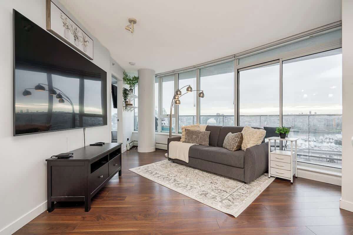 A modern living room with a large flat-screen TV, a grey sofa, a patterned rug, and a city view through large windows.
