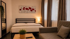 Habitación de hotel con cama, sofá, mesita de noche con planta y control remoto, un cuadro en la pared y una ventana con cortinas.