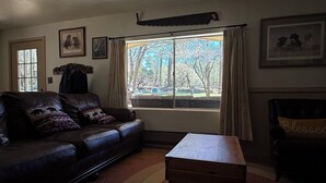A living room with a leather sofa, a wooden chest, and a window with a view of trees. - Serene 2-bedroom cabin with nestled in the pines. (Pine)